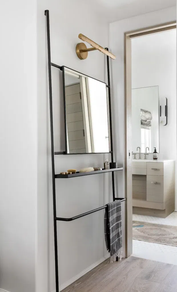 A bright, refined bathroom interior featuring a slim black metal leaning-style storage and towel rail with an integrated rectangular mirror and shelf. A brass wall-mounted light sits above it. Through the open doorway, a white vanity with brass tapware, a large mirror, pale patterned floor tiles, and a neutral rug are visible. The foreground has pale timber-look flooring and white walls.