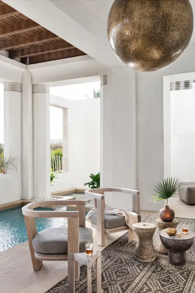 Bright, refined indoor-outdoor lounge beside a small turquoise pool, framed by white plaster columns and walls. The space features pale timber armchairs with upholstered cushions, a patterned dark rug, sculptural side tables, potted plants, and a large perforated bronze pendant light. Exposed dark timber ceiling beams add warmth and architectural interest, while the open courtyard beyond reinforces the airy resort-like atmosphere.