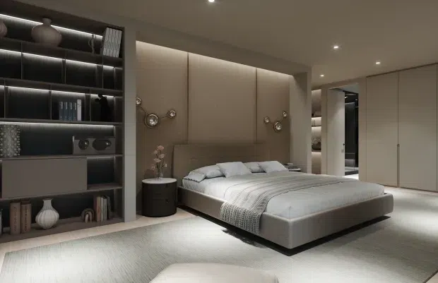 A sophisticated contemporary bedroom with a low-profile upholstered platform bed, soft grey bedding, a textured area rug, and integrated warm LED lighting. Full-height upholstered wall panels form the bedhead, while dark open shelving with decorative objects and books occupies one side. Built-in pale cabinetry and a lit wardrobe or dressing area are visible on the opposite side. The room has a restrained neutral palette, clean lines, and a softly illuminated, luxurious atmosphere.