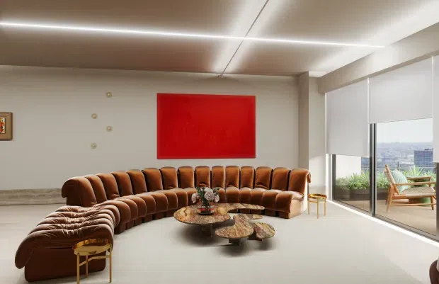 A spacious, refined living room with a large curved tufted brown sectional sofa arranged around an organic sculptural coffee table. The room has pale walls, a light floor, full-height glazing with roller blinds, a red rectangular artwork, recessed linear ceiling lighting, and views toward a cityscape and balcony seating outside. Small gold-toned side tables and decorative objects add warmth and contrast.