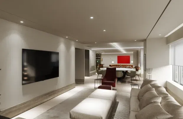 A refined open-plan contemporary interior showing a living room in the foreground with a large wall-mounted television, long upholstered ottoman, and pale sectional sofa beneath expansive windows. A dining area with a large table and upholstered chairs is visible beyond, connected by a linear open layout. The space has a low, smooth ceiling with recessed downlights, warm neutral finishes, pale flooring, and a vivid red artwork or feature panel at the far end.