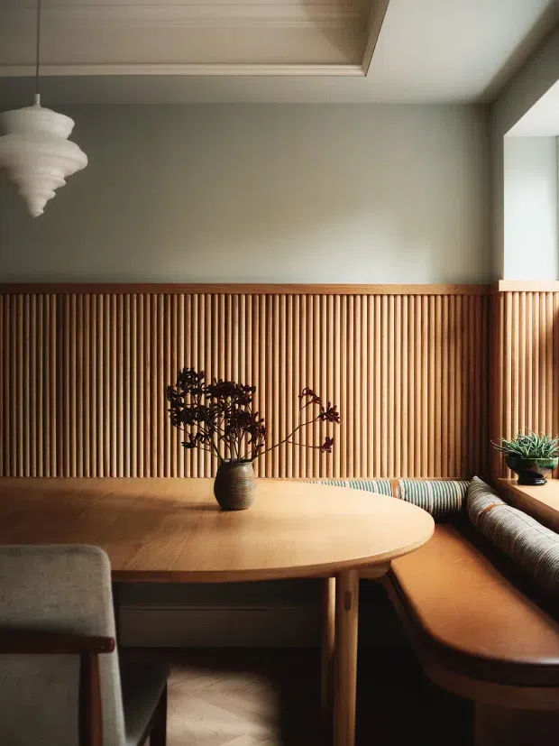 Warm, intimate dining nook with a built-in bench seat wrapping around a rounded timber dining table. The room features vertical timber slat wall panelling, muted taupe plaster walls, a timber-lined recessed ceiling, a sculptural pendant light, and integrated window-side joinery with a potted plant. The styling is natural, refined, and strongly influenced by mid-century forms.