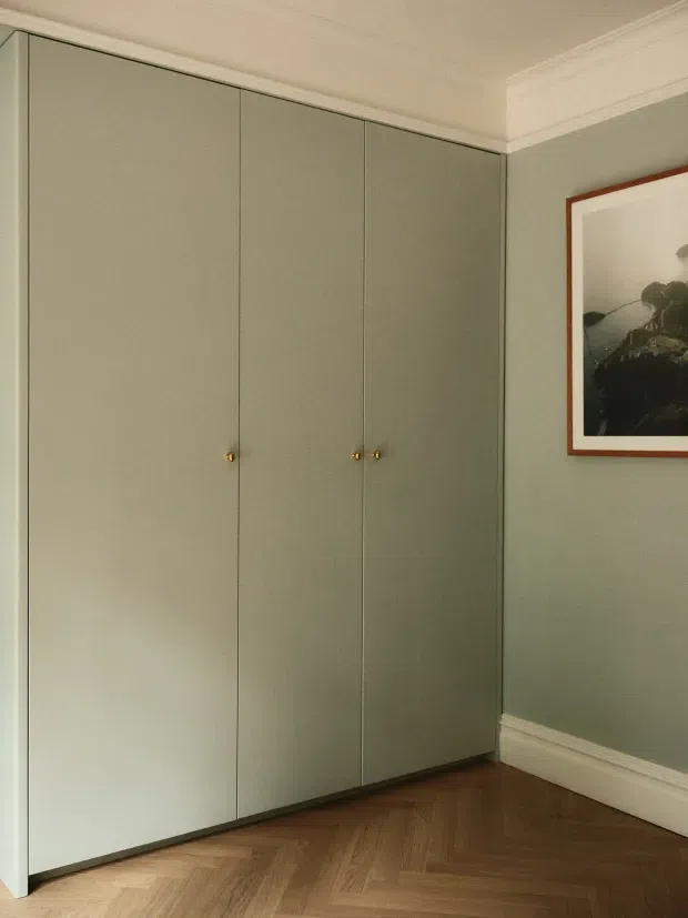 A quiet interior corner with a full-height built-in wardrobe in muted sage-grey cabinetry, small brass knobs, and a classic painted wall with decorative cornices and skirting. The floor is warm herringbone timber, and a framed artwork hangs on the adjacent wall. The composition highlights the fitted storage and refined, understated finishes.