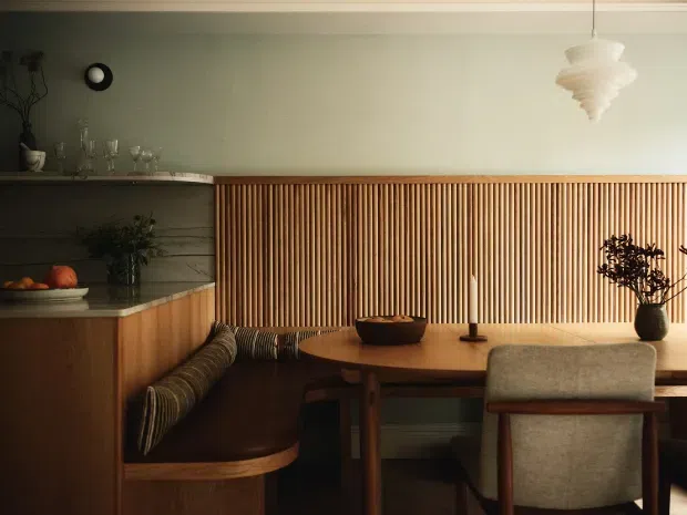 Warm, intimate dining nook with a built-in timber banquette, round wooden dining table, upholstered dining chairs, and a full-height vertical timber slat feature wall. A compact kitchen or serving area with dark cabinetry and stone benchtop is visible to the left. The space is softly lit by a sculptural pendant and styled with ceramics, glassware, and greenery.