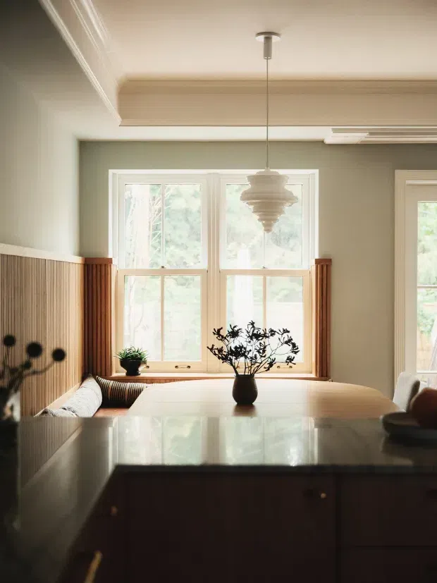 Warm, softly lit dining area viewed from a kitchen island or benchtop. A large timber dining table sits in front of tall white-framed windows, with a built-in banquette along a vertically slatted timber wall. The room features muted sage-green walls, detailed ceiling mouldings, a sculptural pendant light, and a dark stone or engineered-stone kitchen benchtop in the foreground.