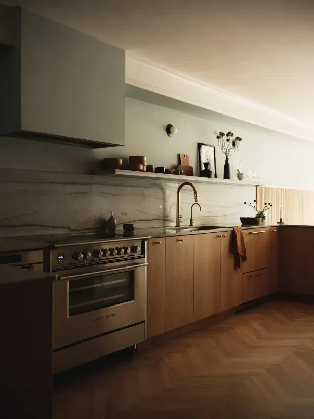 A warm, refined kitchen with a stainless-steel range cooker, dark stone benchtops, timber cabinetry, and a large marble-look backsplash. The kitchen features a sculptural brass gooseneck tap, open display shelving, decorative objects, a concealed rangehood, and parquet timber flooring. Soft natural light and traditional ceiling cornices contrast with the contemporary joinery.