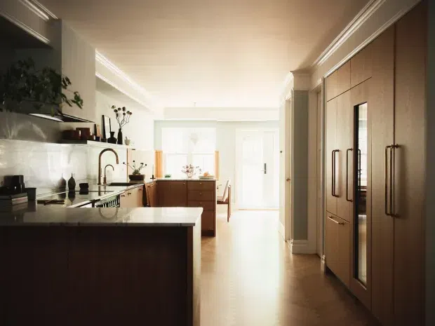 Bright, warm-toned contemporary kitchen viewed along a galley-like layout. The room features dark timber cabinetry, a large light stone island with an undermount sink and curved metal tap, a pale reflective splashback, integrated full-height timber cabinetry with long vertical handles, and classic-style ceiling cornices and wall trims. A dining or living area is visible beyond the kitchen.