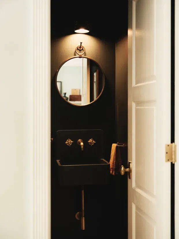 A compact powder room viewed through an open white paneled door. The narrow space has dramatic black walls, a black wall-mounted basin with exposed plumbing, brass/gold taps and fittings, a round framed mirror, and a small wall light above. A warm brown towel hangs beside the basin, while the mirror reflects part of an adjacent light-coloured room.