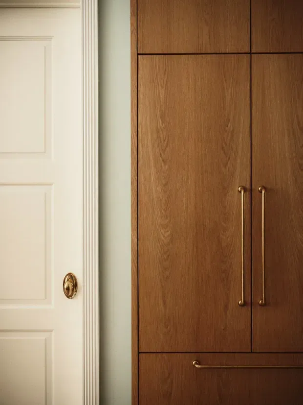 Close-up interior detail showing a traditional cream-painted panelled door with ornate moulded trim and a small brass knob beside built-in dark timber cabinetry. The cabinetry has clean rectangular panels and long slender brass handles, creating a refined blend of classic architectural detailing and warm contemporary joinery.