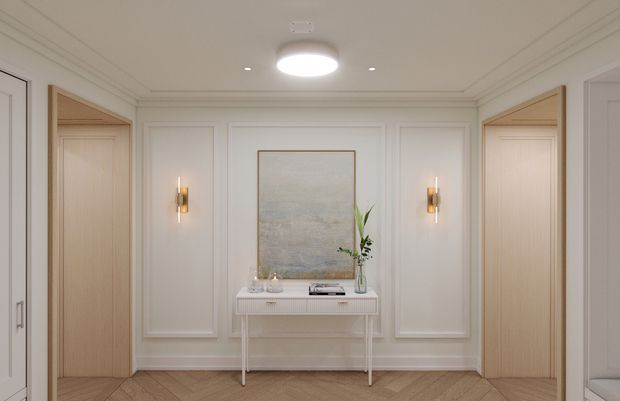 Elegant, symmetrical residential hallway or entry foyer with white painted wall panelling, light timber doorways, a slim console table, framed artwork, paired brass wall sconces, and a central flush-mount ceiling light. The light timber floor is laid in a herringbone pattern, creating a refined and softly warm traditional interior.