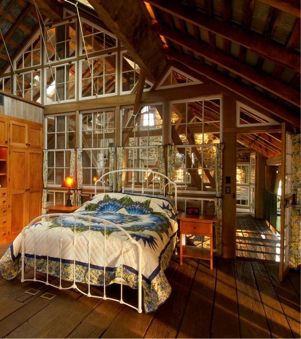 A rustic loft-style bedroom with a white metal four-poster bed, blue patterned bedding, warm timber flooring and extensive timber framing. The room features dramatic exposed roof structure, clerestory-like glazing, large multi-pane windows, mirrored or reflective panels, built-in timber cabinetry, and a small bedside table. Warm natural light creates a cozy, lodge-like atmosphere.