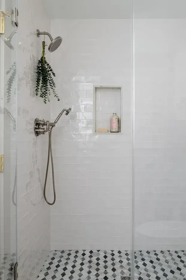 A bright, compact walk-in shower with glossy white subway-tile walls, a patterned black, white and grey mosaic floor, brushed metal shower fittings, a recessed tiled niche, and a clear frameless glass screen. A hanging plant adds a small natural accent.
