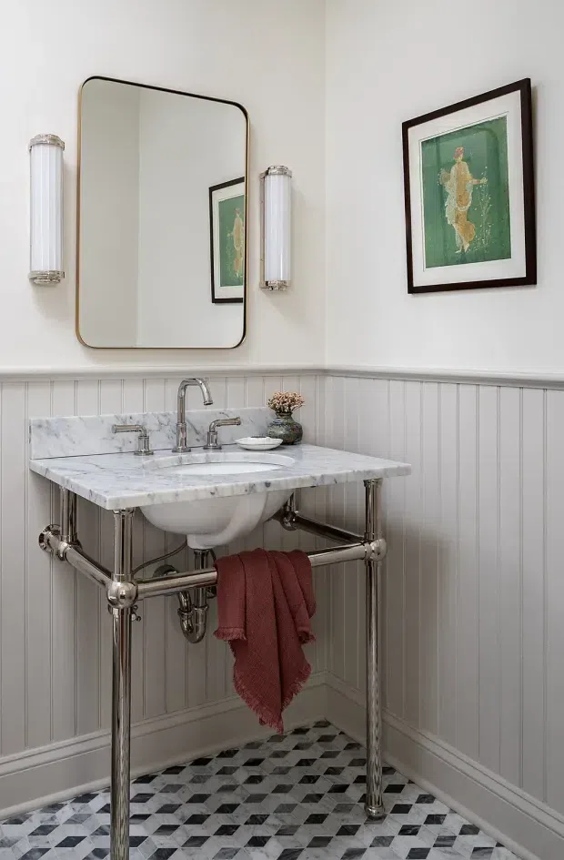 A refined traditional bathroom with a marble console vanity, exposed polished metal plumbing and legs, a white ceramic basin, grey beadboard wall panelling, and a geometric black-and-white tiled floor. A rounded rectangular mirror, flanking wall sconces, and framed artwork add a classic decorative character.