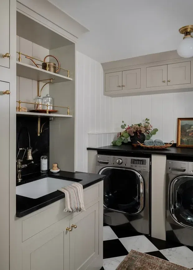 A refined laundry room with pale grey built-in cabinetry, black stone benchtops, a deep utility sink, brass hardware and fittings, open display shelving, and stacked side-by-side stainless-steel washer and dryer appliances. White vertical wall panelling, a black-and-white checkerboard floor, and classic decorative styling create a traditional, polished look.