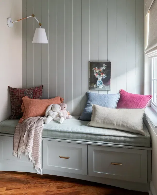 A cozy built-in window seat with drawers, upholstered pale blue-green cushioning, and layered cushions in muted coral, blue, pink, and cream. The seat is set against vertical grey-green wall panelling, with a brass articulated wall light, framed floral artwork, a knitted throw, and a plush toy. Warm timber flooring and abundant natural light create a comfortable reading nook.