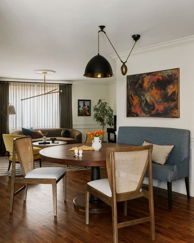 A refined open-plan living and dining room with mid-century-inspired furnishings. A round dark timber dining table is surrounded by upholstered timber dining chairs, while a blue upholstered sofa sits behind it. The room features polished dark timber flooring, white walls with decorative wall moulding, full-height sheer and dark drapery, sculptural black and brass pendant lighting, and a large abstract artwork in warm orange, red, and charcoal tones.