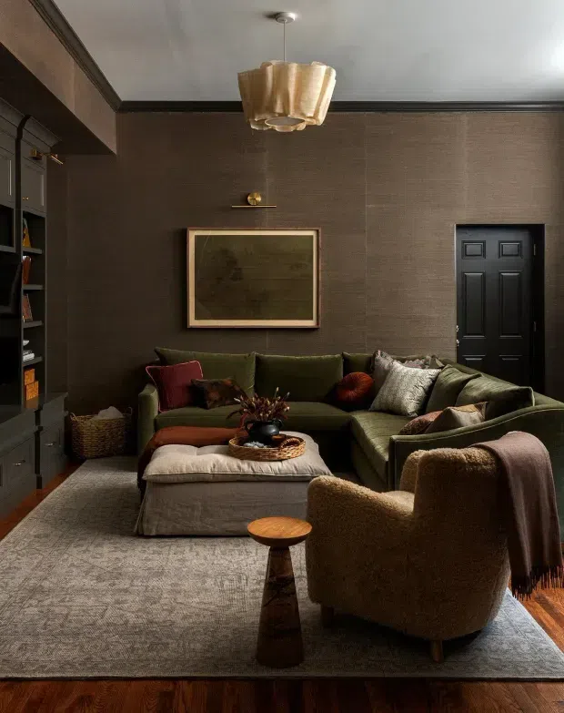 A richly layered, moody living room with dark textured walls, black trim, built-in shelving, a black paneled door, and warm wood flooring. An olive-green sectional sofa, upholstered armchair, ottoman, cushions, area rug, side table, and sculptural pendant light create a cosy, refined seating area.