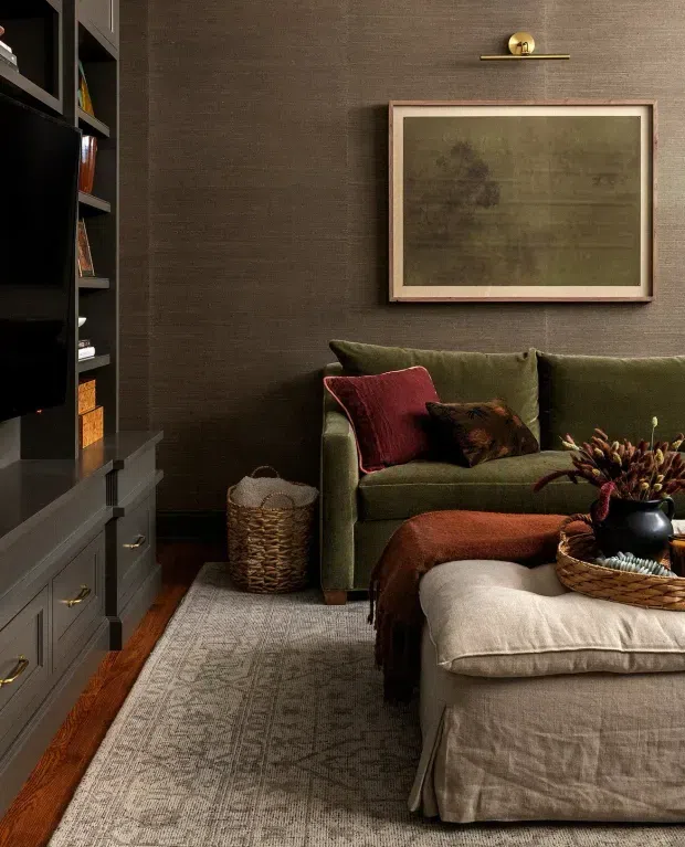 A cozy, moody living room featuring a deep olive-green sofa with layered cushions, a large upholstered ottoman, dark built-in media cabinetry, textured dark brown wallcovering, warm timber flooring, and a neutral patterned area rug. A framed abstract artwork and brass picture light add refined accents, while woven baskets and dried botanical stems provide natural texture.