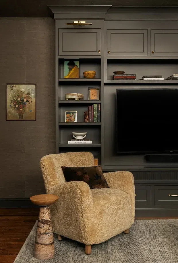 A warm, moody living room corner with a boucle lounge chair and patterned cushion positioned in front of dark built-in media cabinetry. The cabinetry includes paneled doors, open display shelving, brass-toned hardware, a wall-mounted television and concealed storage. Dark textured wallcovering, timber flooring, a textured area rug, framed artwork and a small sculptural side table complete the refined, cosy setting.