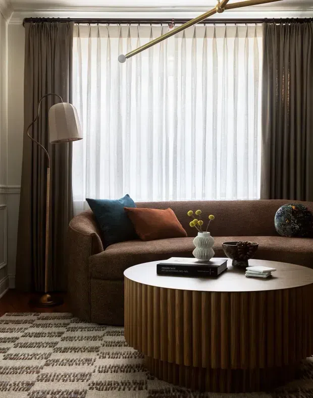 A refined living room with a curved brown upholstered sofa positioned in front of a large window with sheer white curtains and dark drapes. A sculptural floor lamp, round white coffee table with a fluted dark-wood base, patterned rug, decorative cushions, vase, books, and art object create a warm, layered interior. Traditional wall mouldings contrast with contemporary furniture and lighting.