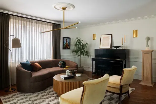 A refined living room with a curved brown sofa, two cream upholstered armchairs, a round fluted timber coffee table, black upright piano, framed artwork, decorative wall panelling, and a sculptural brass-and-white ceiling light. Full-height sheer curtains and dark drapes frame the windows, while a patterned rug and warm timber flooring complete the room.