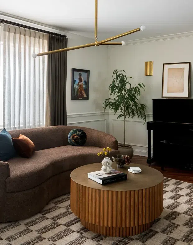 A refined living room with a curved brown upholstered sofa, round fluted timber coffee table, black upright piano, patterned rug, framed artwork, indoor plant, and a sculptural brass-and-white branching pendant light. Full-height sheer and dark drapery frame the window, while decorative wall moulding adds architectural detail.