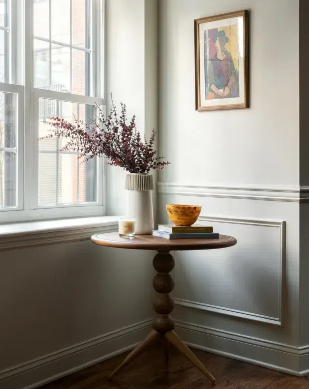 A refined interior vignette in a bright corner beside tall white-framed windows. A round dark timber pedestal console table displays a ceramic vase with burgundy branches, a candle, books, and a wooden bowl. The walls feature white decorative panel moulding and a framed abstract artwork, while dark timber flooring adds contrast. The composition feels elegant, traditional, and carefully styled.