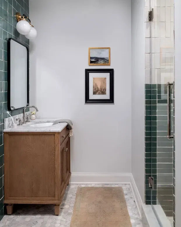 A compact, traditionally styled bathroom with a timber vanity, white marble countertop and integrated basin, black-framed rounded mirror, brass wall light, and framed artwork. Dark green and white tiled walls surround the vanity and shower, while the floor features small pale mosaic tiles and a neutral bath mat.