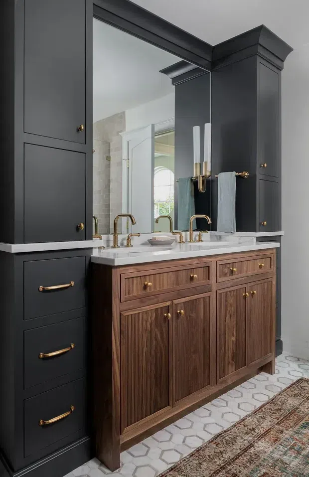 A refined bathroom vanity area featuring a large framed wall mirror, a double-sink marble or engineered-stone countertop, warm brass faucets and hardware, and richly grained fluted timber cabinetry. Tall charcoal built-in cabinetry flanks the vanity, while pale geometric floor tiles and a patterned rug add contrast. A tiled shower or wet area is visible in the mirror reflection.
