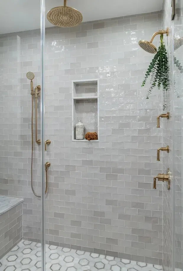 A bright, refined walk-in shower with glossy pale grey subway-tile walls, patterned hexagonal floor tiles, brass shower fixtures, a built-in recessed niche, and a frameless glass enclosure. A hanging green plant adds a natural accent.