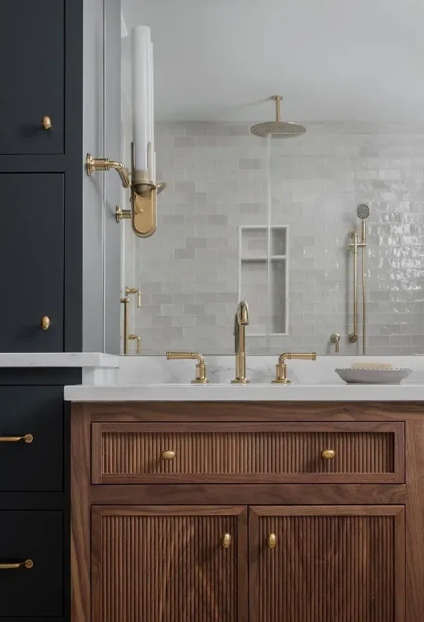 Close-up view of an elegant bathroom vanity with dark navy built-in cabinetry at left, a warm fluted timber vanity with brass hardware, white benchtop, and polished brass tapware. The bathroom features glossy white rectangular wall tiles, a built-in shower niche, overhead rain shower, and handheld shower fittings. The composition is refined and warmly lit, with a classic contemporary aesthetic.
