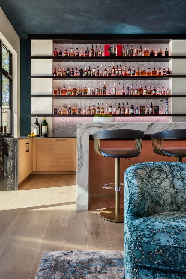 A sophisticated residential home bar with a backlit display wall lined with liquor bottles, dark open shelving, a veined marble bar front, timber cabinetry, and two upholstered brass-based bar stools. The room has wide pale timber flooring, a dark textured ceiling, a patterned blue armchair in the foreground, and strong natural light from a window on the left.