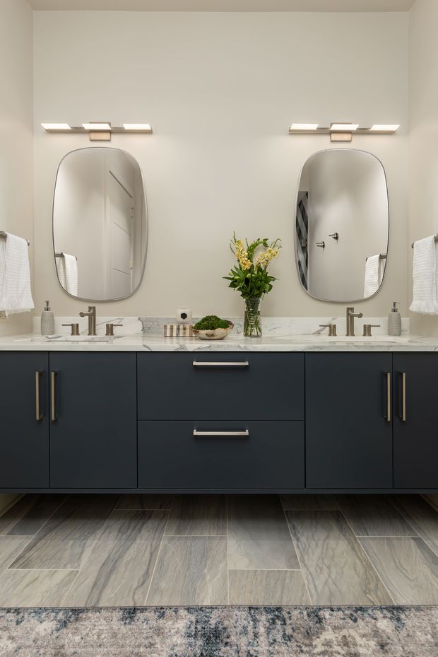 A refined double-vanity bathroom with a long dark navy or charcoal floating cabinet, white marble-look benchtop, two integrated basins, brushed metal faucets, rounded rectangular mirrors, and warm horizontal wall lights. Large grey stone-look floor tiles and a patterned rug add texture, while white towels and a small floral arrangement soften the scheme.