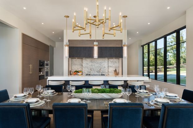 A refined open-plan dining and kitchen space with a large dark timber dining table set for entertaining, upholstered navy dining chairs, and a long white kitchen island behind it. The kitchen features dark wood cabinetry, a pale stone island and backsplash, integrated appliances, and a dark vertical-ribbed feature panel. A striking brass and glass multi-arm chandelier hangs above the dining table, while expansive black-framed windows provide views to a pool and landscaped outdoors.