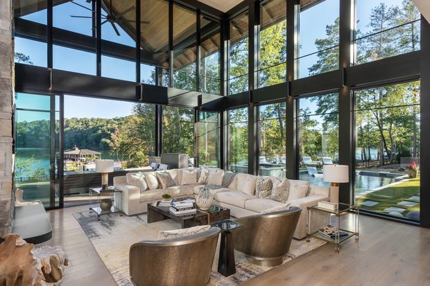 A luxurious double-height living room with expansive floor-to-ceiling black-framed glazing, exposed timber ceiling panels, and views to a landscaped pool terrace and wooded setting. The room contains a large cream sectional sofa, sculptural upholstered lounge chairs, glass coffee tables, floor lamps, and a pale timber floor. Strong indoor–outdoor connection and abundant natural light create an airy, refined atmosphere.
