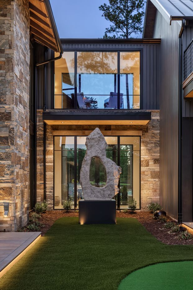 A sophisticated modern courtyard framed by multi-level wings of a residence. The architecture combines stacked natural stone walls, dark vertical metal cladding, extensive black-framed glazing, and exposed timber eaves. A sculptural stone artwork stands on a dark plinth in the centre of a landscaped artificial-grass courtyard, with subtle integrated lighting along the pathway and a curved green putting or lawn area in the foreground.