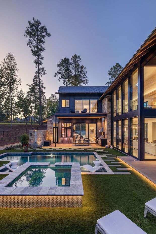 Twilight exterior view of a contemporary multi-level residence with extensive floor-to-ceiling glazing, dark metal cladding and trim, warm timber soffits, and stone accents. The landscaped backyard features a swimming pool with an integrated raised spa, paved coping, artificial lawn, stepping stones, and outdoor lounge furniture. Interior living areas are visible through the illuminated glazing.