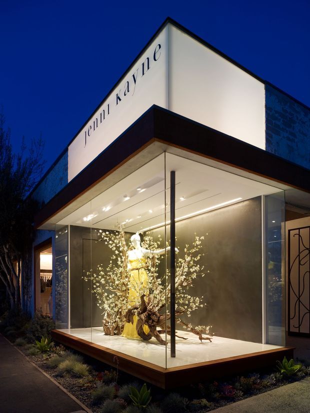 Twilight exterior view of a contemporary retail or commercial storefront with a sharply projecting flat canopy, illuminated full-height glass display windows, and a prominent illuminated sign reading “Cella Kayne.” A mannequin in a yellow outfit is arranged within a sculptural floral display, while dark cladding, clean geometric lines, and integrated lighting define the facade.