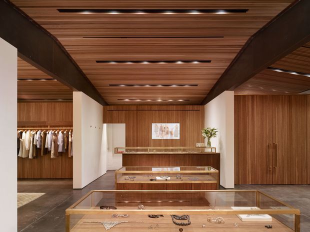 A refined luxury retail showroom displaying jewellery in illuminated glass-topped timber cases. The interior features extensive warm vertical timber wall panelling and a slatted timber ceiling with integrated recessed lighting, framed by substantial dark exposed beams. Clothing is displayed in an open wardrobe area at the left, while tall timber cabinetry occupies the right side. The space has a warm, polished, gallery-like atmosphere.