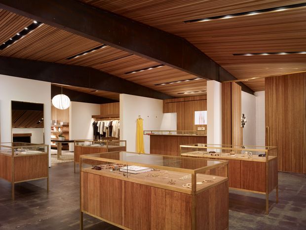 A refined retail showroom with warm timber display cases, glass-topped counters, wood wall panelling, and a slatted timber ceiling. Dark exposed structural beams frame the space, while recessed linear lighting and a spherical pendant provide illumination. Clothing racks, jewellery displays, mirrors, and a rear service counter are visible, creating a boutique-like commercial interior.