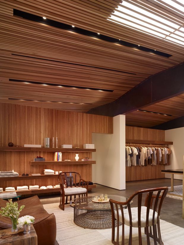 A refined retail showroom or boutique interior with extensive warm timber slat panelling and a matching slatted ceiling. Open clothing racks and built-in timber display shelving line the rear and side walls. Dark structural beams, recessed linear lighting, a concrete-look floor, lounge seating, timber dining-style chairs, a low wire coffee table, and a leather sofa create a warm, sophisticated atmosphere.