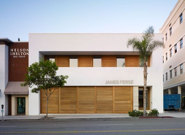 Street-level photograph of a contemporary two-storey commercial building with a clean white rendered facade, deep horizontal openings, and extensive timber louvred screening over the windows and ground-floor frontage. The building is identified as James Perse, with an adjacent Nelson Shelton Real Estate office and neighbouring multi-storey commercial development. A palm tree and street frontage are visible.