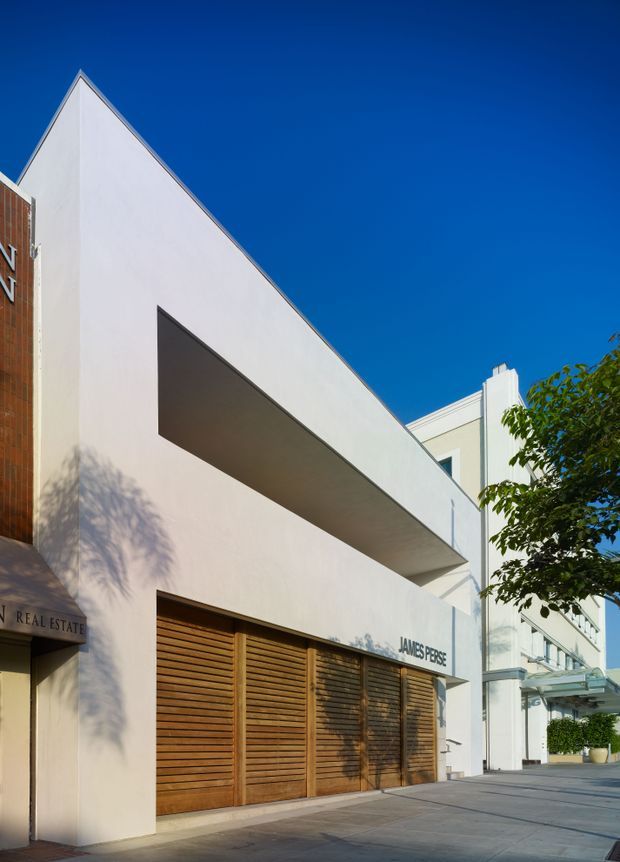 Street-level view of a contemporary urban commercial building facade. The building has a sharply sloping triangular parapet, smooth pale rendered walls, a long recessed horizontal opening, timber louvred garage or storefront doors, and glazed entry elements. Signage is visible on the facade, with adjacent buildings and street trees in the background.