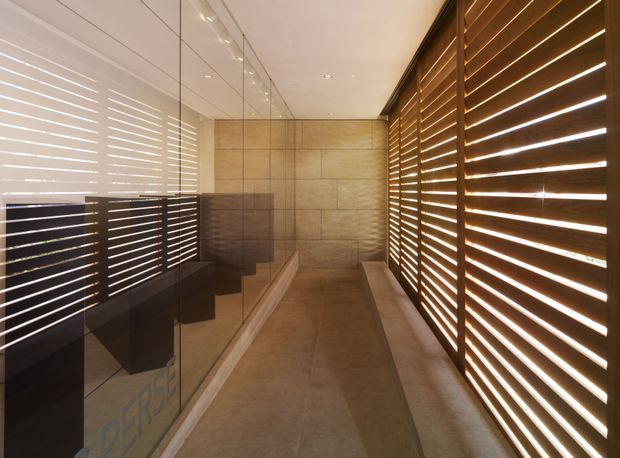 A narrow, refined interior passage or entry area with a long glass wall on the left and dark timber horizontal louvres on the right filtering strong linear daylight. The floor and rear wall are finished in warm beige stone or large-format tile, with a pale flush ceiling and minimal detailing. Reflections in the glazing suggest dark built-in joinery or cabinetry beyond the glass. The space has a warm, contemporary architectural character with strong light-and-shadow contrast.
