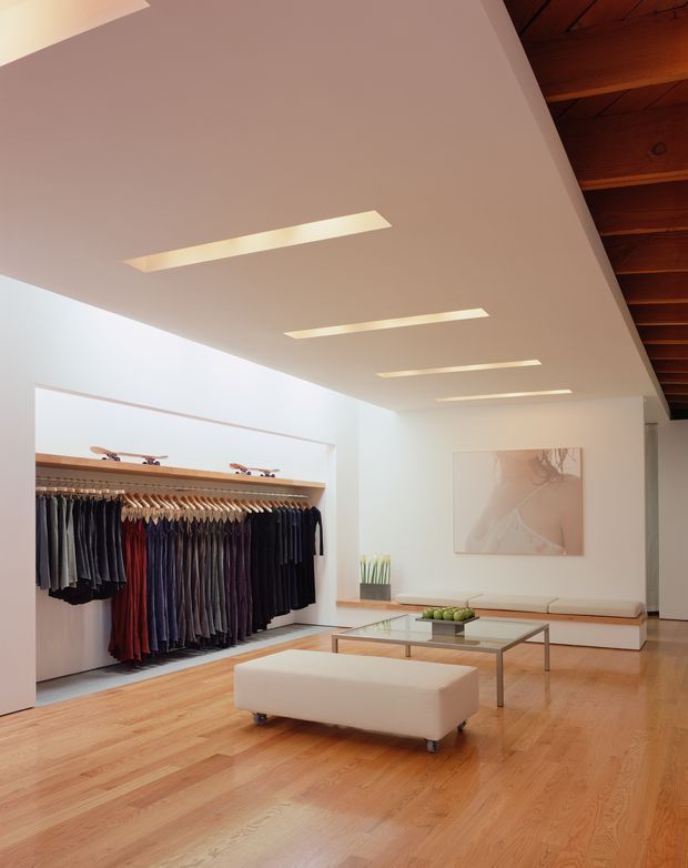 Bright, minimalist clothing boutique with garments displayed on a long wall-mounted rail, polished timber flooring, recessed linear ceiling lights, a low white display bench, glass coffee tables, and a warm exposed timber ceiling section. The space has clean white walls and understated contemporary retail styling.