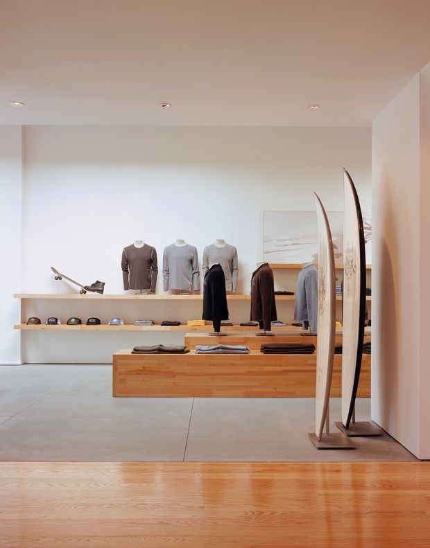 A minimalist contemporary clothing and surfwear retail interior with pale plaster walls, polished concrete flooring, warm timber display counters and shelving, recessed ceiling lights, mannequins and neatly arranged garments. Tall surfboards form a sculptural display near the entrance, while caps and accessories line the rear shelving. The space has a clean, understated, warm-toned showroom aesthetic.