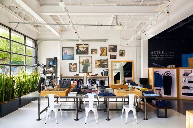 Bright industrial-style retail showroom with a high exposed white ceiling, large black-framed glazed garage-style windows, clothing displays, wall-mounted artwork, and a central arrangement of long tables and white chairs. The space combines apparel retail with a workshop-like bicycle display area and abundant indoor planting.