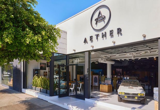 Street-level exterior photograph of an AETHER retail storefront in bright daylight. The modern white facade has prominent raised signage and circular logo, dark metal-framed full-height glazing, and a wide open garage-style retail entrance. Merchandise and a display vehicle are visible inside, with a small planted frontage and leafy street tree.