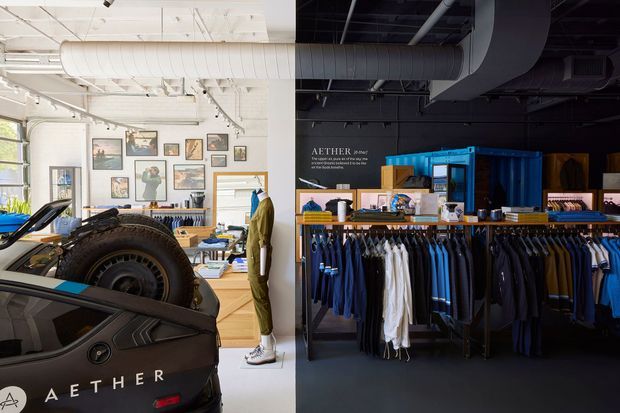 A split-view commercial retail interior for AETHER, combining a bright automotive and fashion showroom. The left side has white walls and ceiling services, framed artwork, a mannequin, clothing displays, and a large rugged vehicle or vehicle component in the foreground. The right side is a darker, industrial-style retail area with exposed ductwork, black walls and ceiling, blue shelving and display fixtures, and neatly arranged clothing racks.