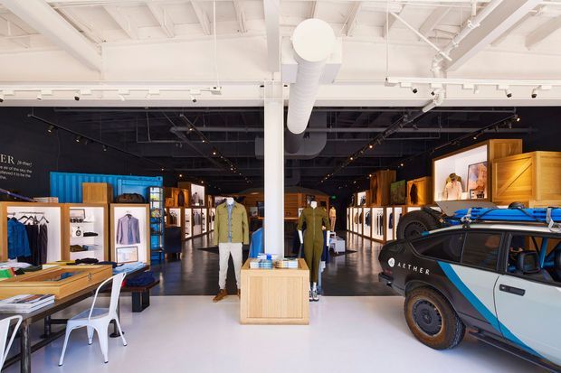 A bright contemporary retail showroom with a high exposed white-painted ceiling, prominent ductwork, track lighting, and polished concrete flooring. Clothing displays, shelving, timber display plinths, worktables, and mannequins fill the space, while a branded off-road vehicle occupies the foreground. The interior combines a clean white front area with a darker rear display zone and industrial-style services.